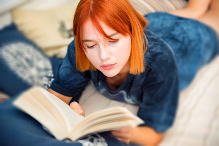 Pretty red-haired teenage girl reads book in her room